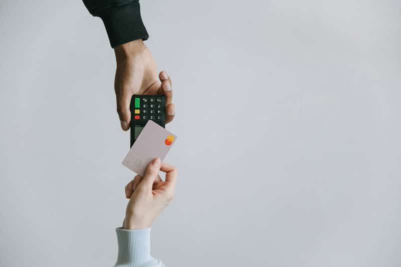 A person's hand tapping their card to pay while another person's hand holds the card machine out, solid grey background.