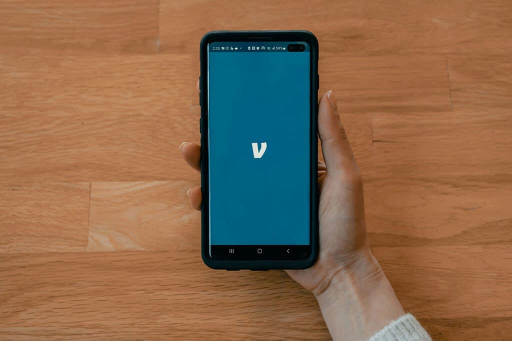 A person holding a smartphone showing a blue screen background with the Venmo "V" logo in the center against a wooden table.