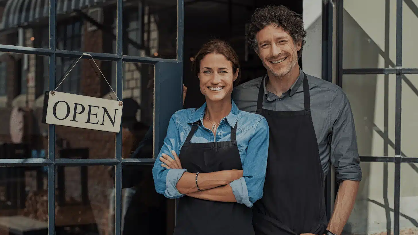 two smiling business owners standing next to open sign