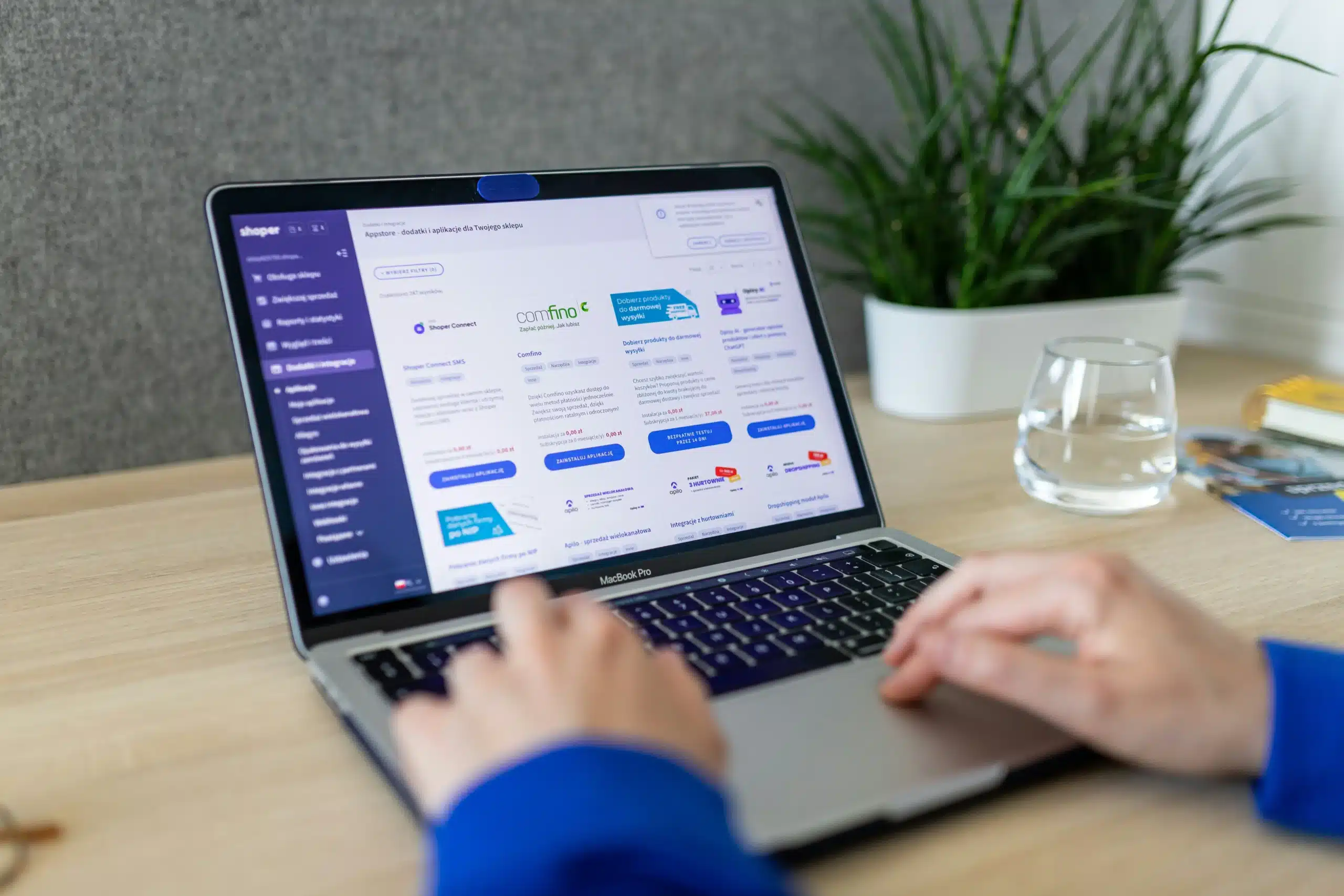 Hands typing on a MacBook Pro displaying an e-commerce platform integrations dashboard on a desk, with a potted plant and a glass of water in the background.