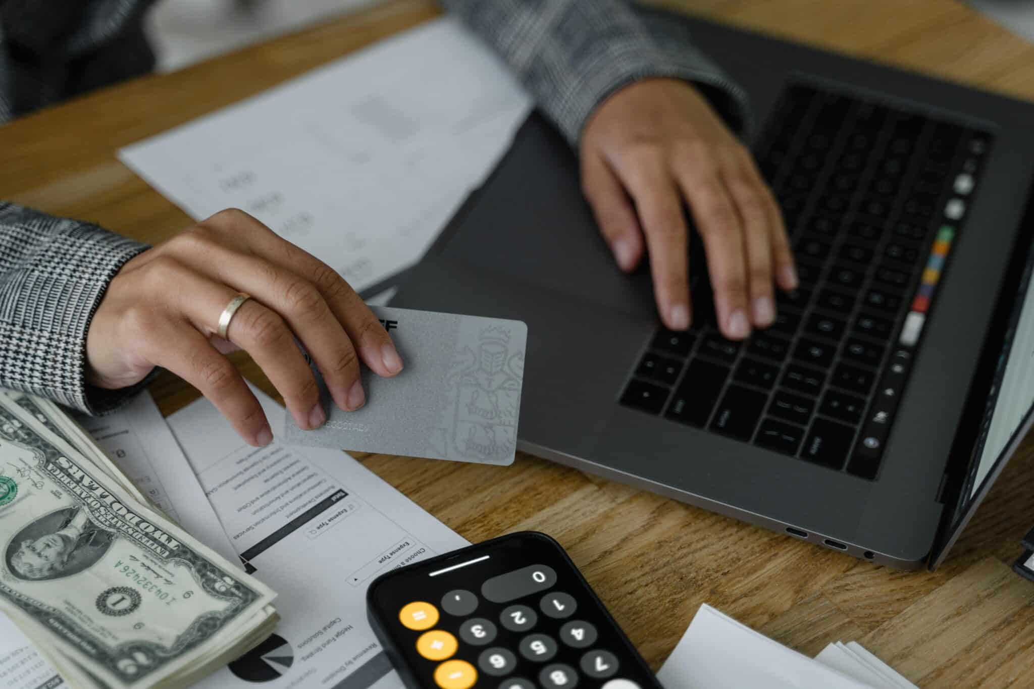 a hand holding a credit card and using a laptop on a desk with a calculator, cash, and documents