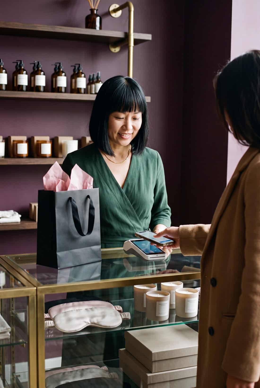 Adult boutique owner at a glass counter as a customer taps a phone on a card terminal to pay